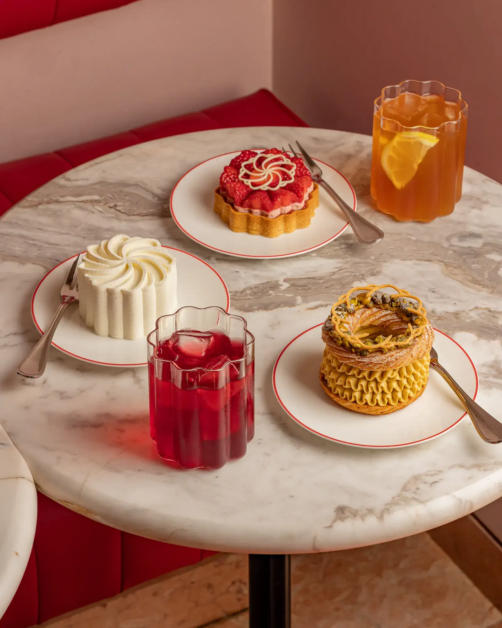Three plated pastries and two drinks on marble café table with red booth seating.