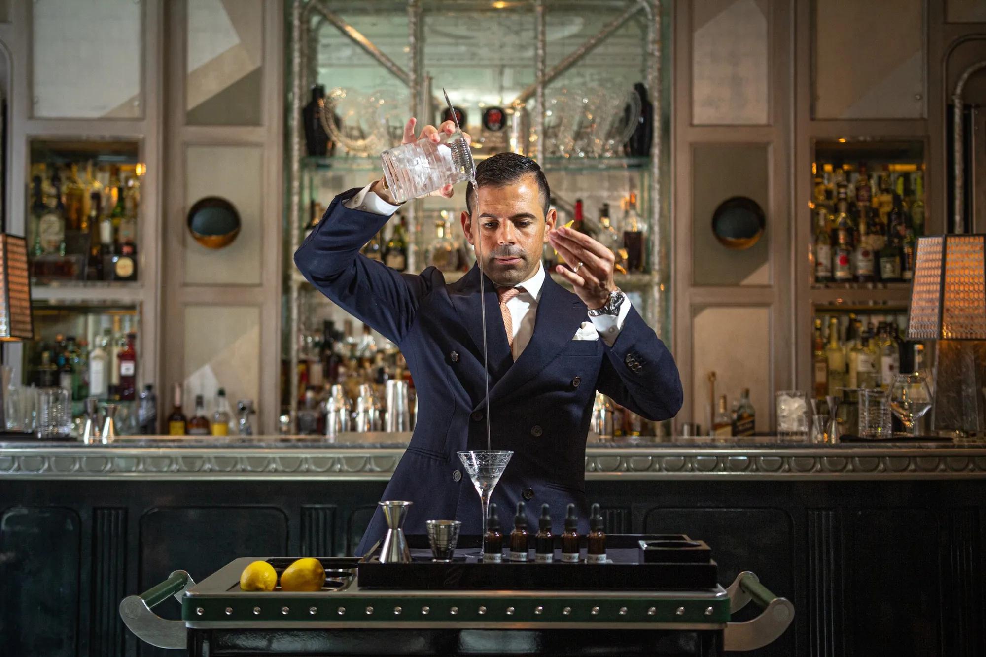 Bartender elegantly pours a martini from height into a glass on a cocktail trolley in a refined bar.