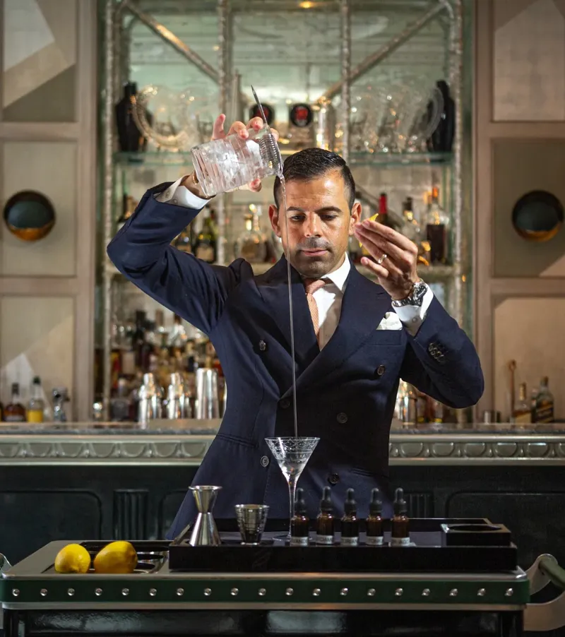 Bartender elegantly pours a martini from height into a glass on a cocktail trolley in a refined bar.