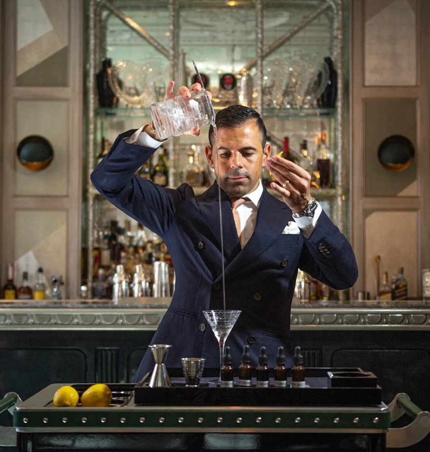 Bartender elegantly pours a martini from height into a glass on a cocktail trolley in a refined bar.