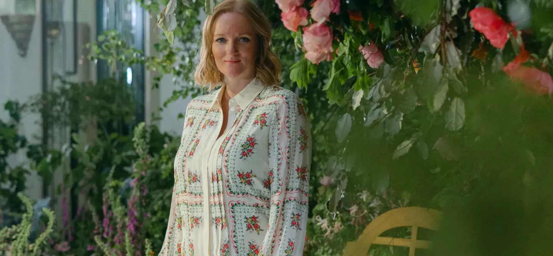 Woman in embroidered white dress standing in lush indoor garden with pink flowers and greenery, smiling at camera.