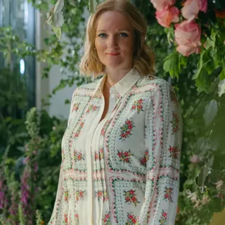 Woman in embroidered white dress standing in lush indoor garden with pink flowers and greenery, smiling at camera.