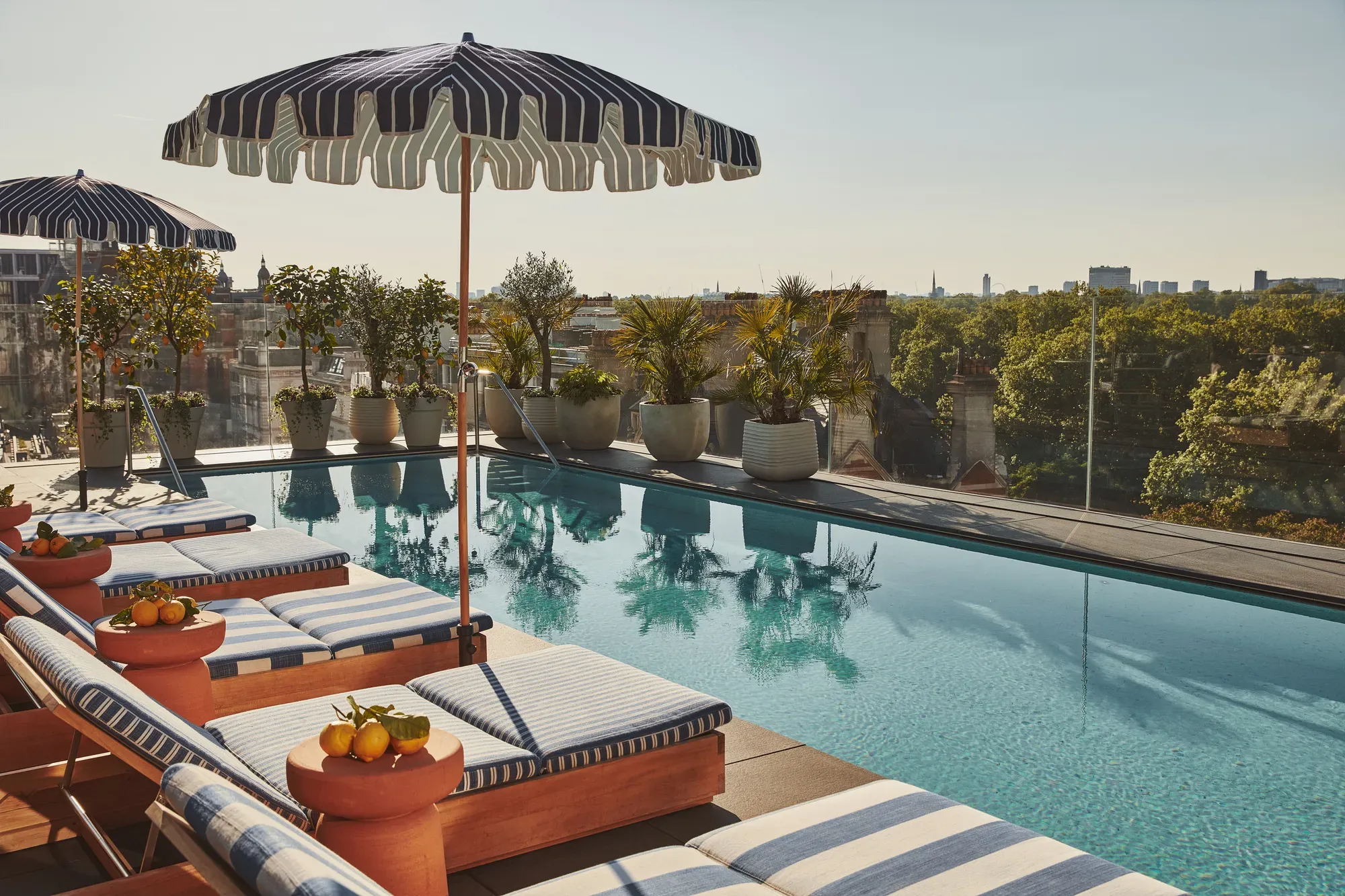 Rooftop pool with striped loungers and umbrellas, overlooking trees and city views under the summer sun.