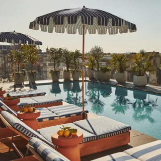 Rooftop pool with striped loungers and umbrellas, overlooking trees and city views under the summer sun.