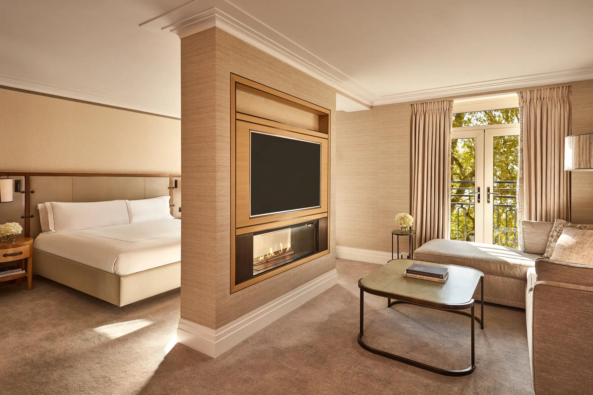 Spacious suite with bed, fireplace, TV, and seating area beside French doors with park views
