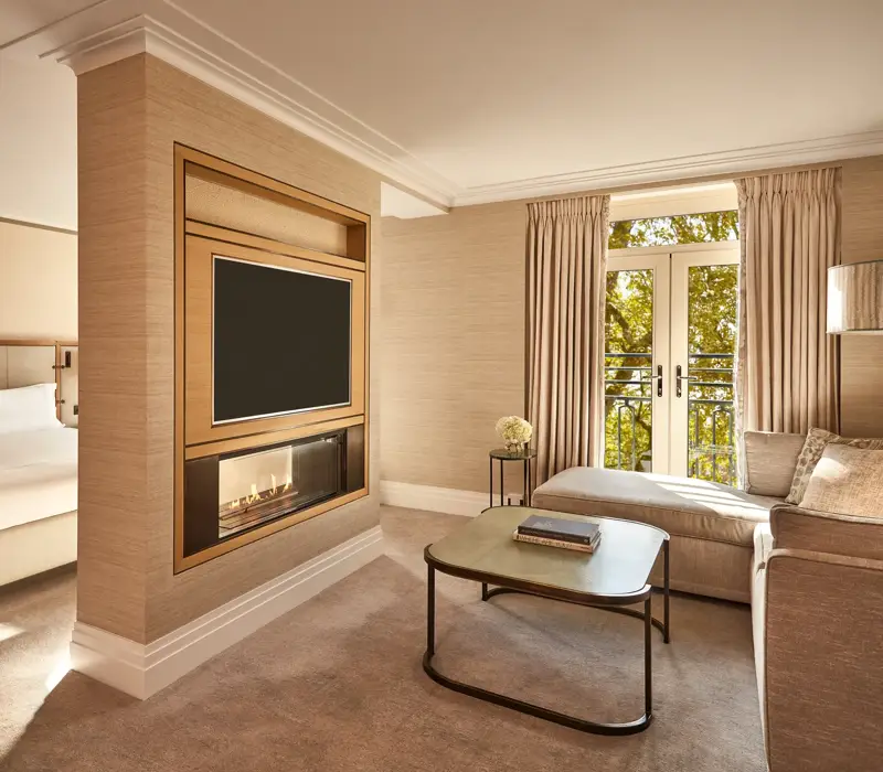 Spacious suite with bed, fireplace, TV, and seating area beside French doors with park views