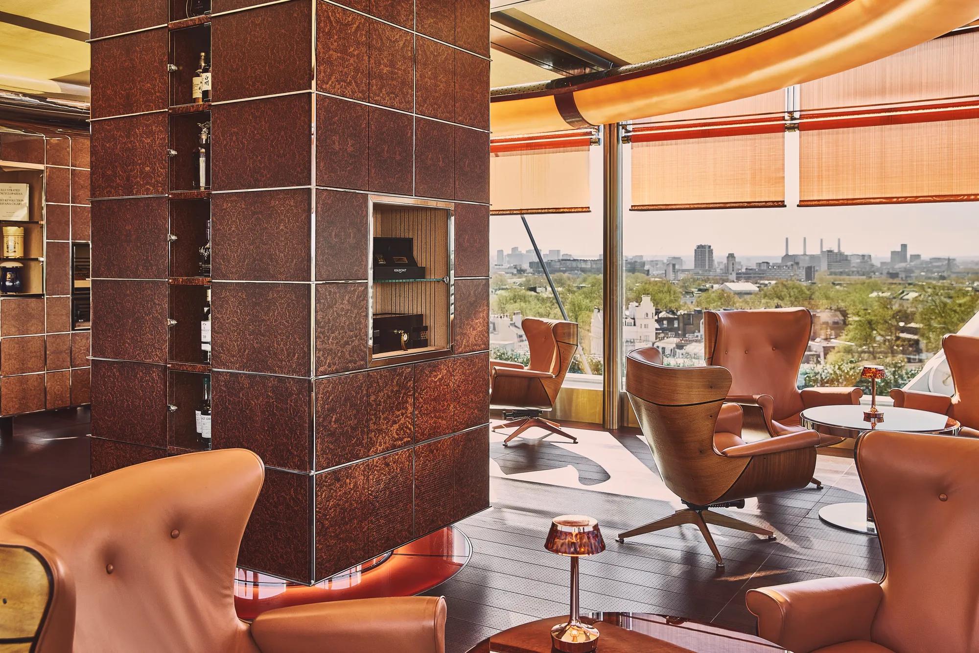 Leather lounge chairs beside a copper-clad fireplace with panoramic city views through floor-to-ceiling windows.