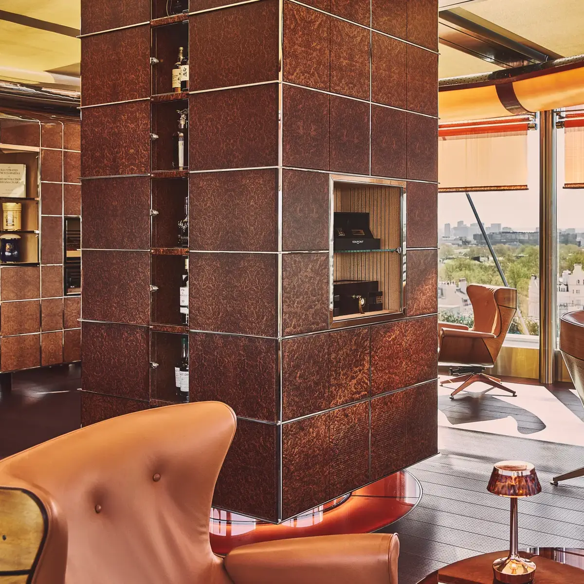 Leather lounge chairs beside a copper-clad fireplace with panoramic city views through floor-to-ceiling windows.