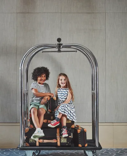 Two children sitting on stacked suitcases atop a hotel luggage trolley, smiling against a neutral interior backdrop.