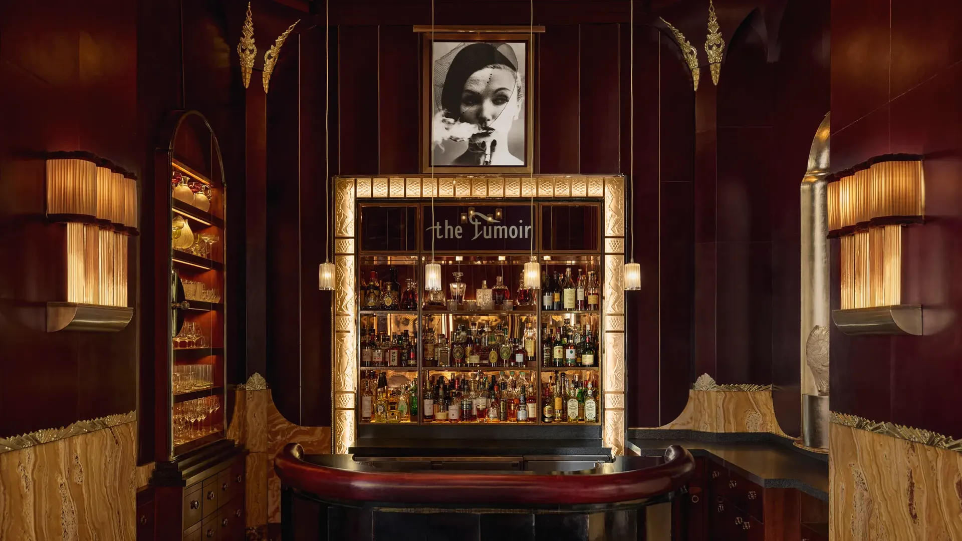 Intimate cocktail bar with rich burgundy and gold tones, featuring a curved leather counter, three stools, and a backlit display of spirits framed by art deco detailing. A black-and-white portrait hangs above the bar, adding to the sophisticated, vintage ambiance.
