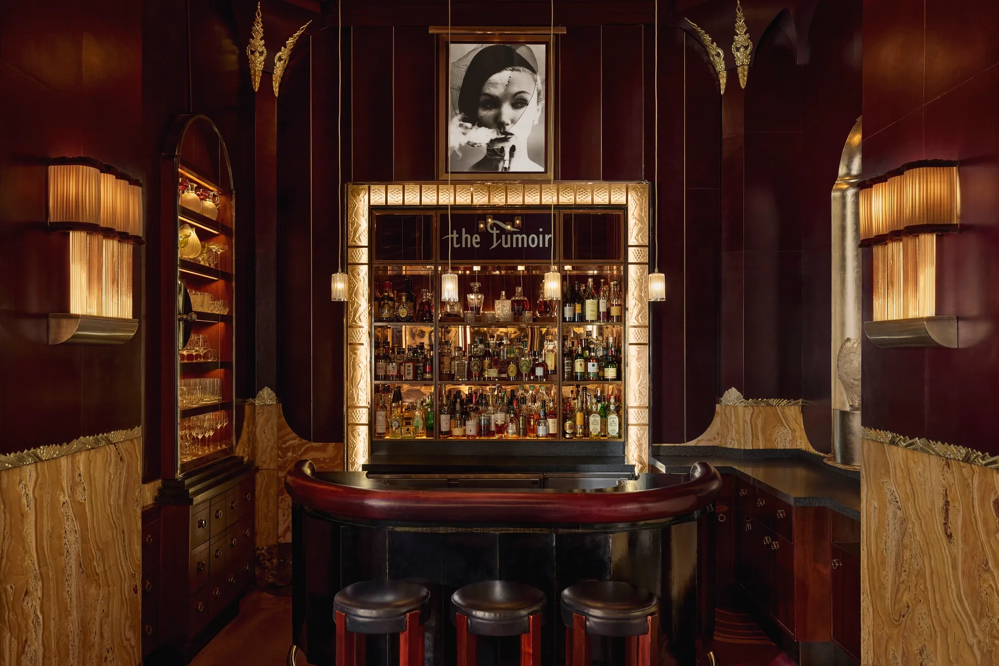 Intimate cocktail bar with rich burgundy and gold tones, featuring a curved leather counter, three stools, and a backlit display of spirits framed by art deco detailing. A black-and-white portrait hangs above the bar, adding to the sophisticated, vintage ambiance.