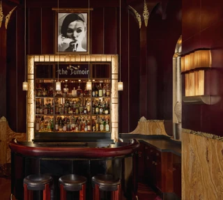 Intimate cocktail bar with rich burgundy and gold tones, featuring a curved leather counter, three stools, and a backlit display of spirits framed by art deco detailing. A black-and-white portrait hangs above the bar, adding to the sophisticated, vintage ambiance.