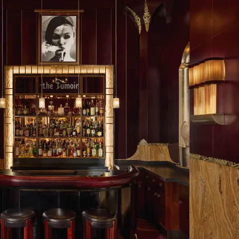 Intimate cocktail bar with rich burgundy and gold tones, featuring a curved leather counter, three stools, and a backlit display of spirits framed by art deco detailing. A black-and-white portrait hangs above the bar, adding to the sophisticated, vintage ambiance.