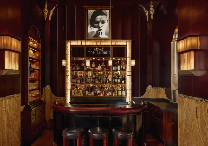 Intimate cocktail bar with rich burgundy and gold tones, featuring a curved leather counter, three stools, and a backlit display of spirits framed by art deco detailing. A black-and-white portrait hangs above the bar, adding to the sophisticated, vintage ambiance.