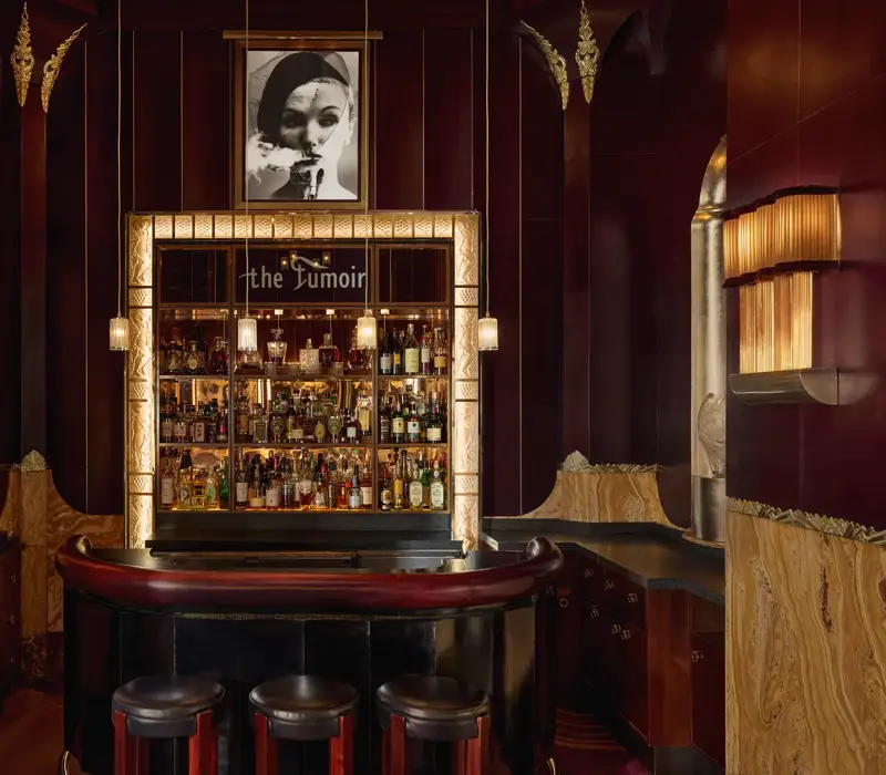 Intimate cocktail bar with rich burgundy and gold tones, featuring a curved leather counter, three stools, and a backlit display of spirits framed by art deco detailing. A black-and-white portrait hangs above the bar, adding to the sophisticated, vintage ambiance.