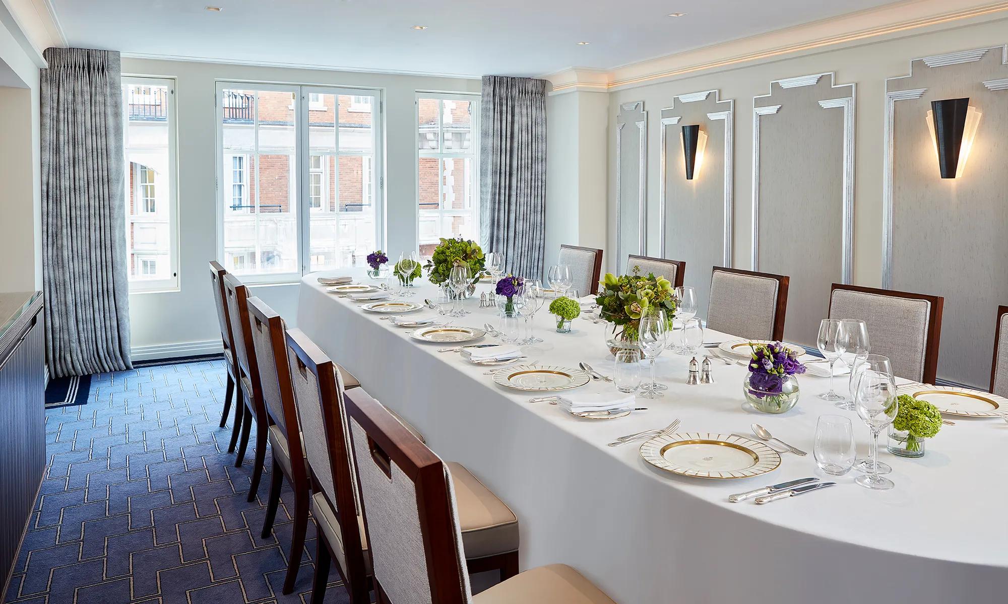 Bright private dining room at Claridge’s with a long oval table set with fine china, glassware, and green and purple floral arrangements beneath tall windows with grey curtains.