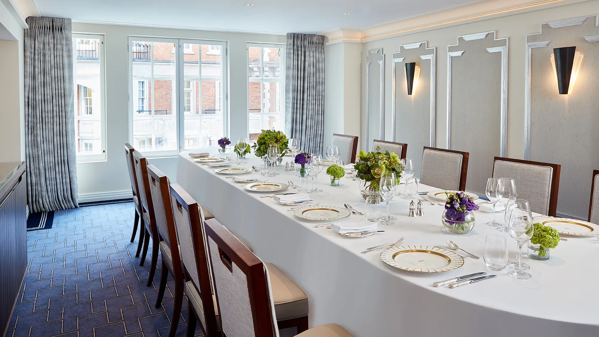Bright private dining room at Claridge’s with a long oval table set with fine china, glassware, and green and purple floral arrangements beneath tall windows with grey curtains.