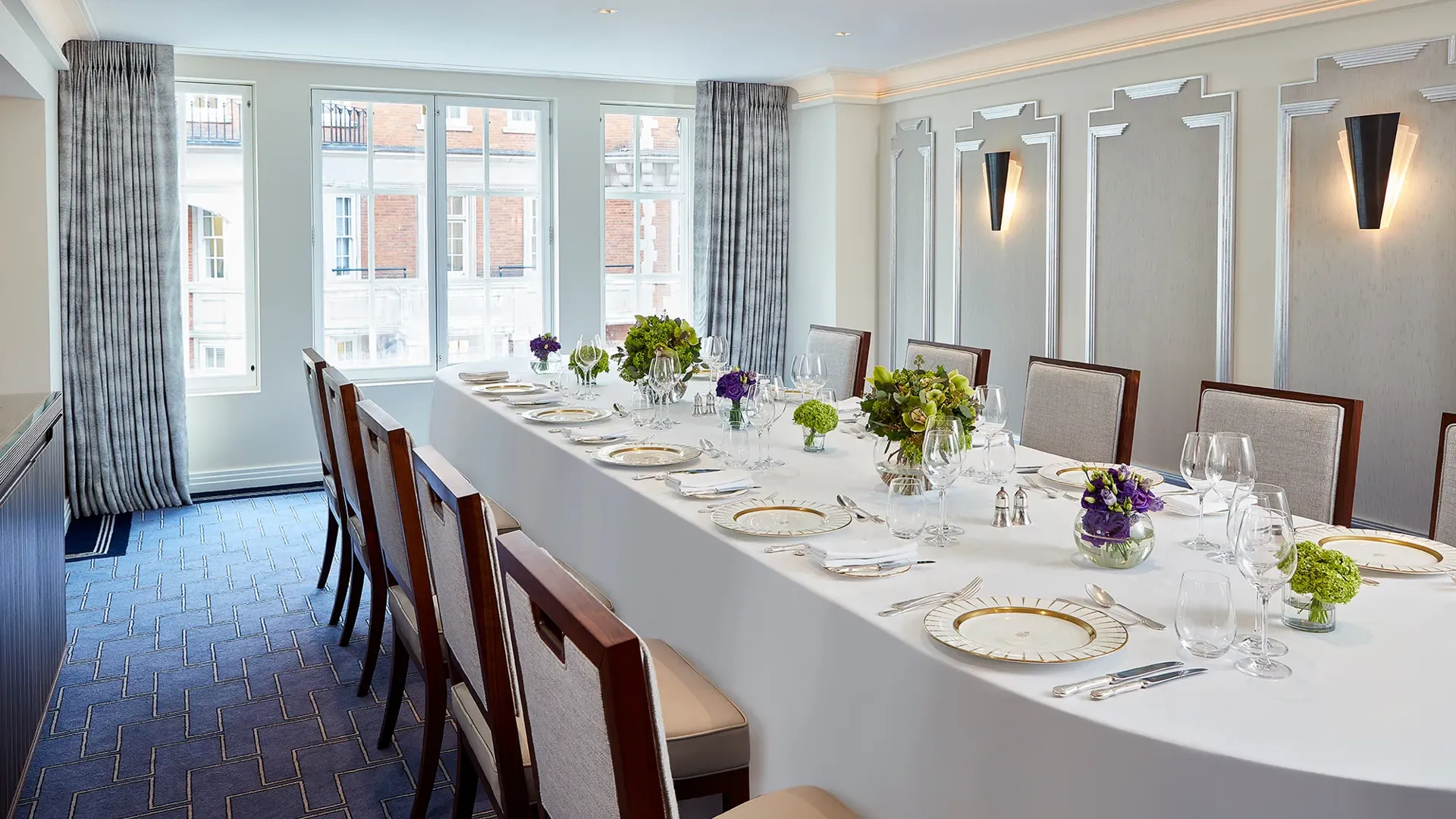Bright private dining room at Claridge’s with a long oval table set with fine china, glassware, and green and purple floral arrangements beneath tall windows with grey curtains.