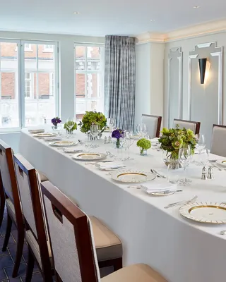 Bright private dining room at Claridge’s with a long oval table set with fine china, glassware, and green and purple floral arrangements beneath tall windows with grey curtains.