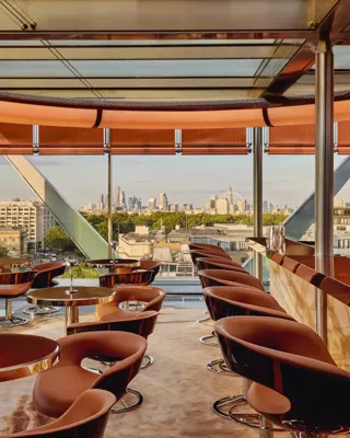 Stylish rooftop bar with caramel leather chairs, marble counter, and panoramic views of London through floor-to-ceiling windows.