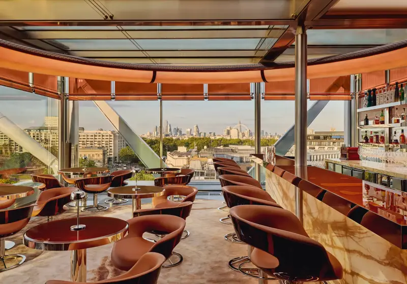 Stylish rooftop bar with caramel leather chairs, marble counter, and panoramic views of London through floor-to-ceiling windows.