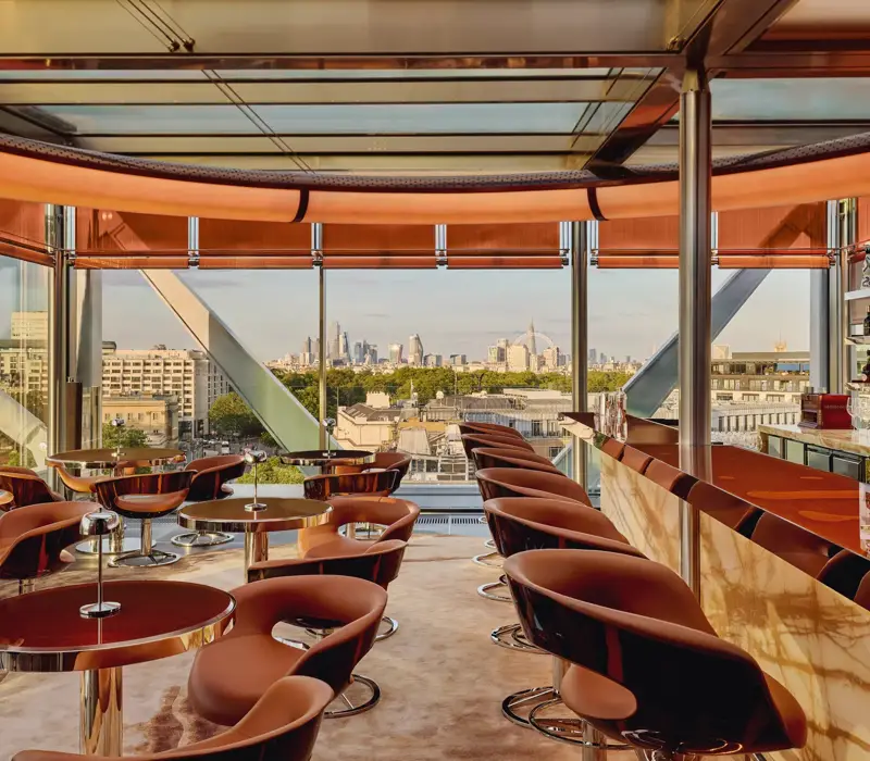 Stylish rooftop bar with caramel leather chairs, marble counter, and panoramic views of London through floor-to-ceiling windows.