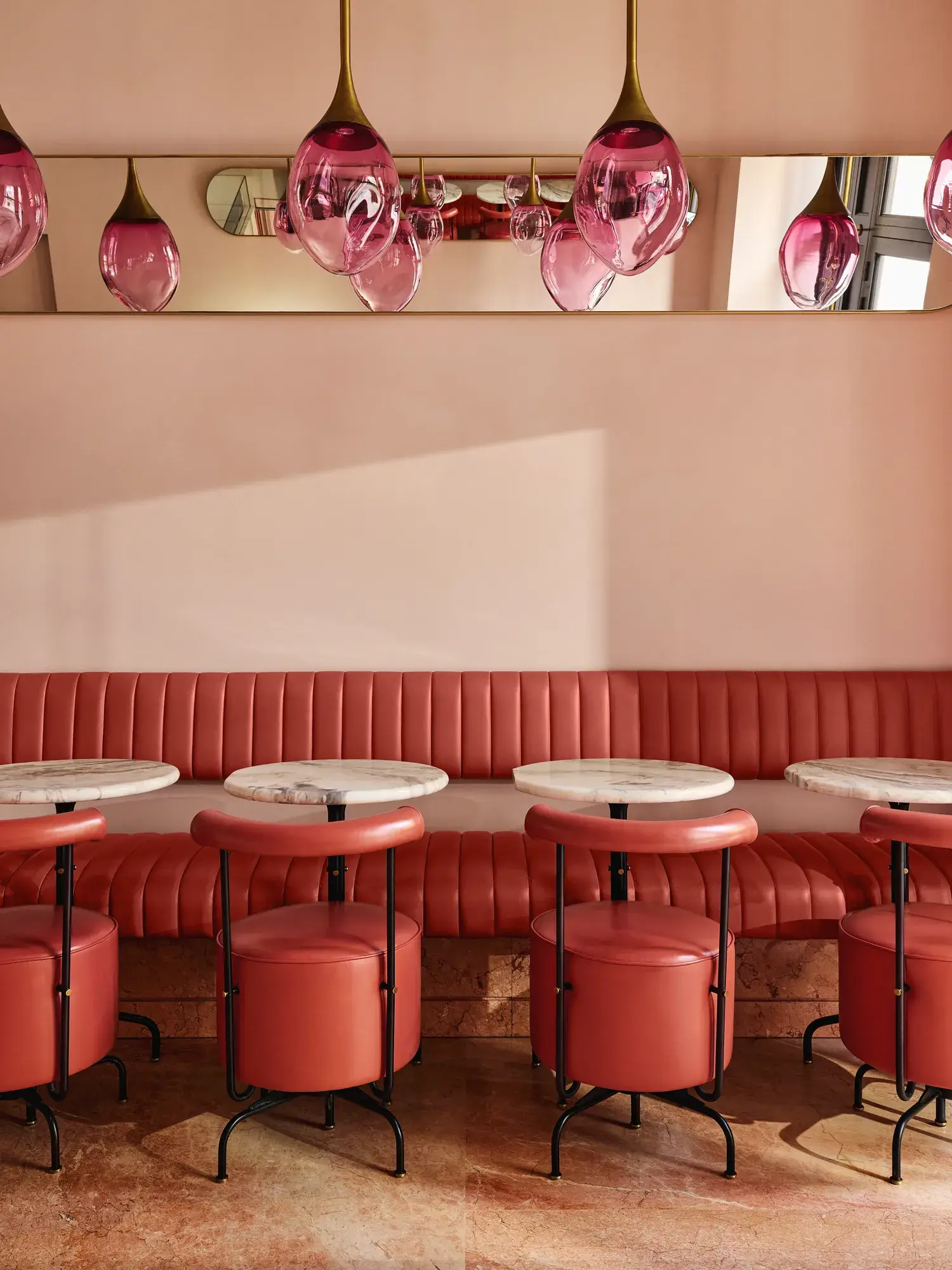 Plush pink banquette seating with round marble tables and pink glass pendant lights in a softly lit café.