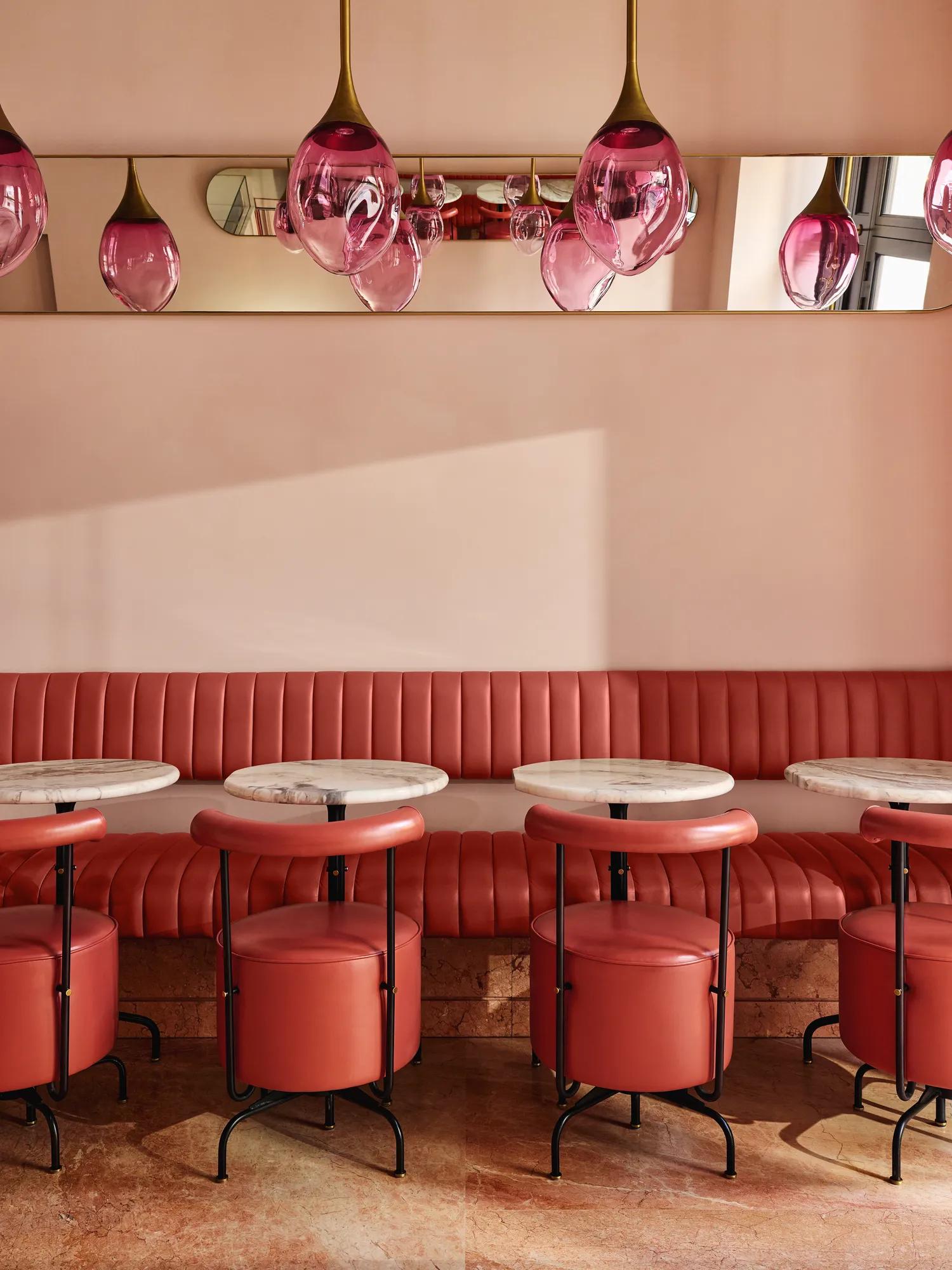 Plush pink banquette seating with round marble tables and pink glass pendant lights in a softly lit café.