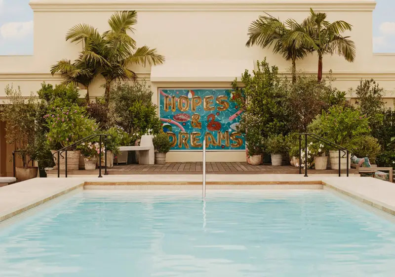 Guest steping from The Maybourne Beverly Hills rooftop pool toward the “Hopes & Dreams” mural, palms and planters behind.