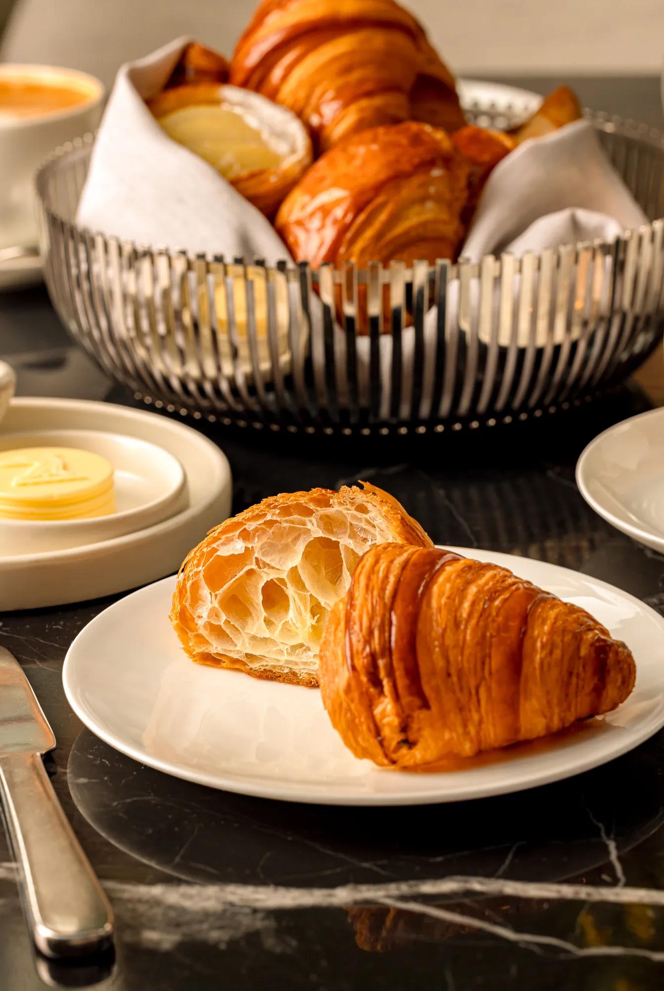 Fresh croissant, halved to show flaky layers, on a white plate with a basket of croissants and butter on a marble table.