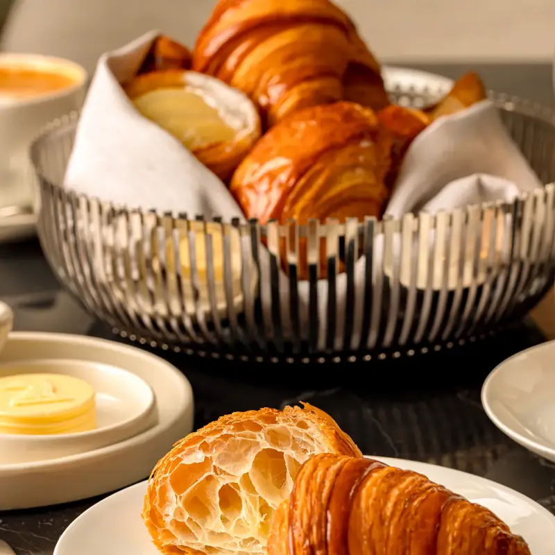 Fresh croissant, halved to show flaky layers, on a white plate with a basket of croissants and butter on a marble table.