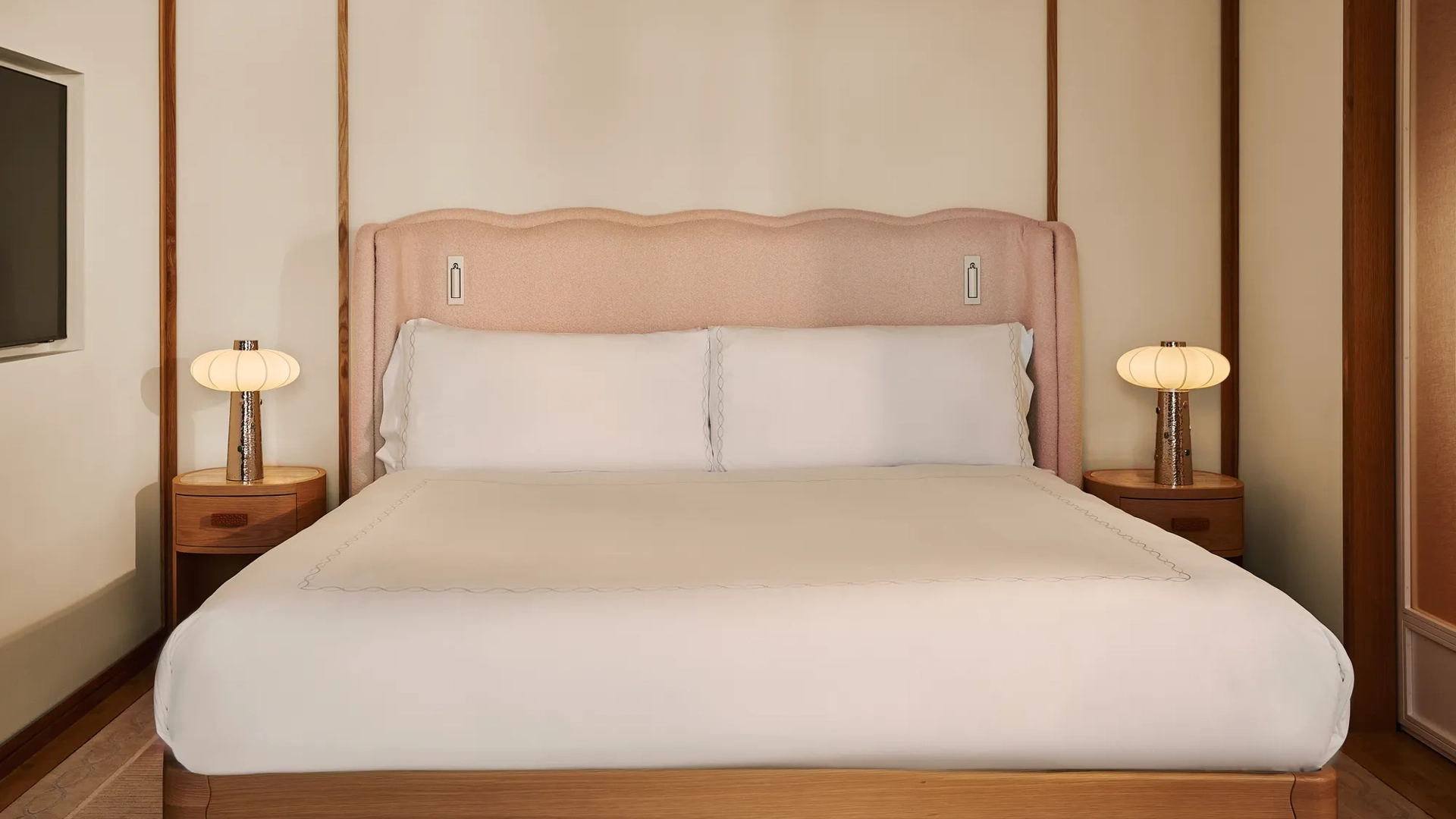 King bed with upholstered pink headboard, white linens, wooden bedside tables with lamps, and paneled walls.