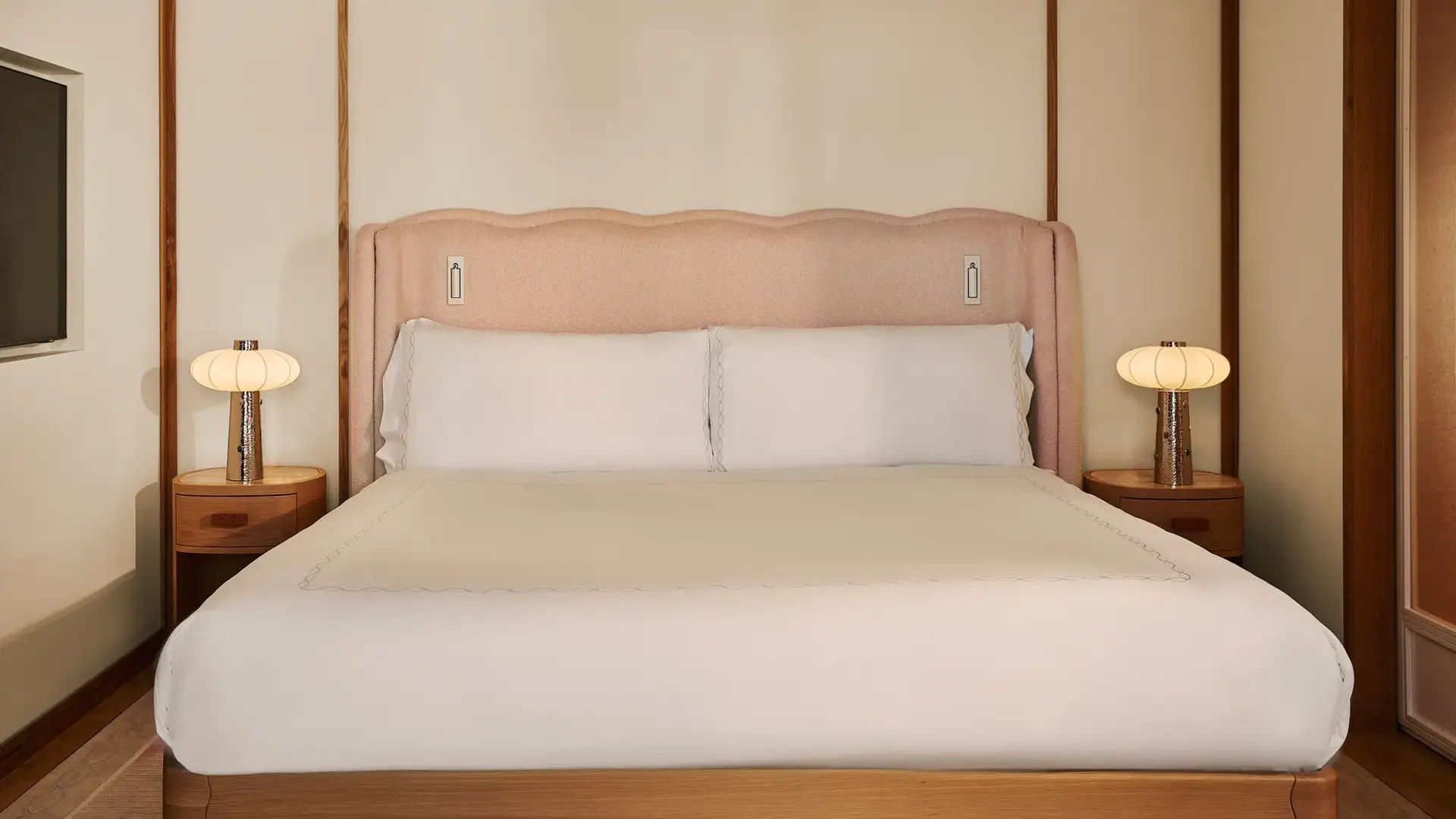 King bed with upholstered pink headboard, white linens, wooden bedside tables with lamps, and paneled walls.