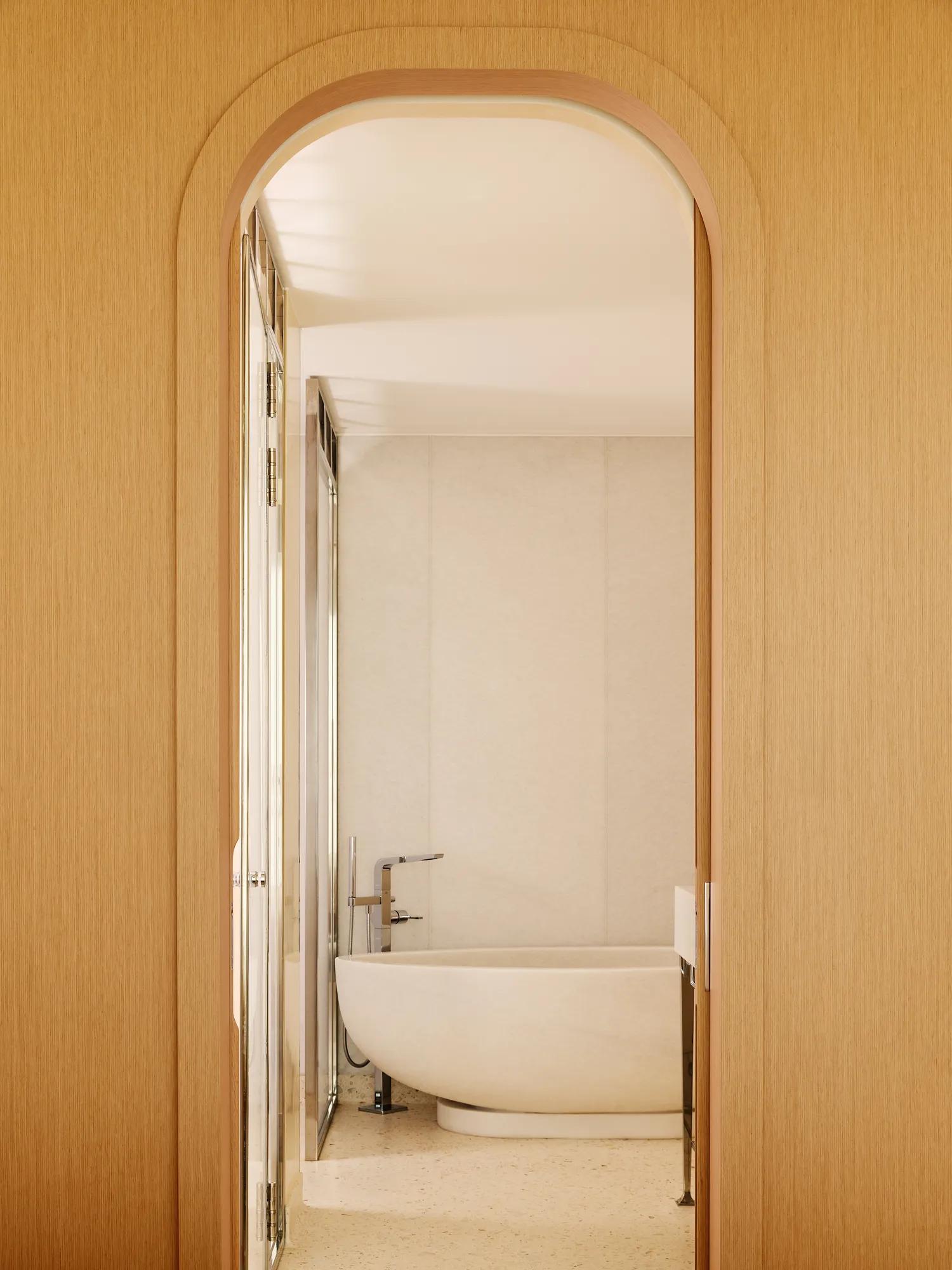 Salle de bain minimaliste avec baignoire ovale autoportante, vue à travers une porte arquée en bois.