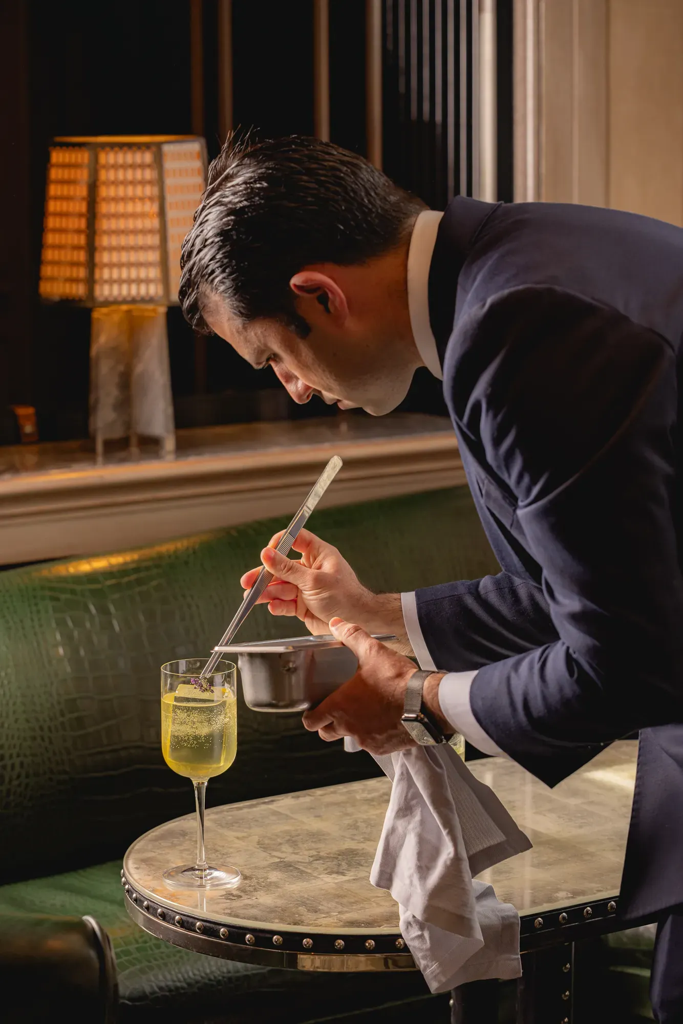 Bartender in a suit carefully preparing a cocktail at a small table in a softly lit bar.