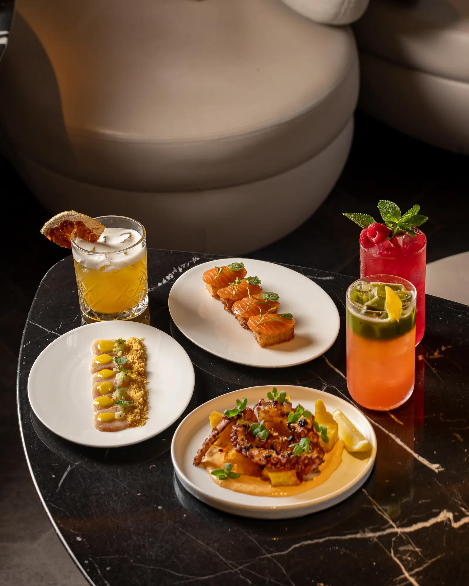 Selection of shared small plates with salmon bites, grilled octopus, dips, and colourful cocktails on a dark marble table.