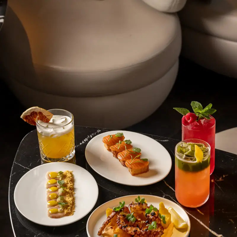 Selection of shared small plates with salmon bites, grilled octopus, dips, and colourful cocktails on a dark marble table.