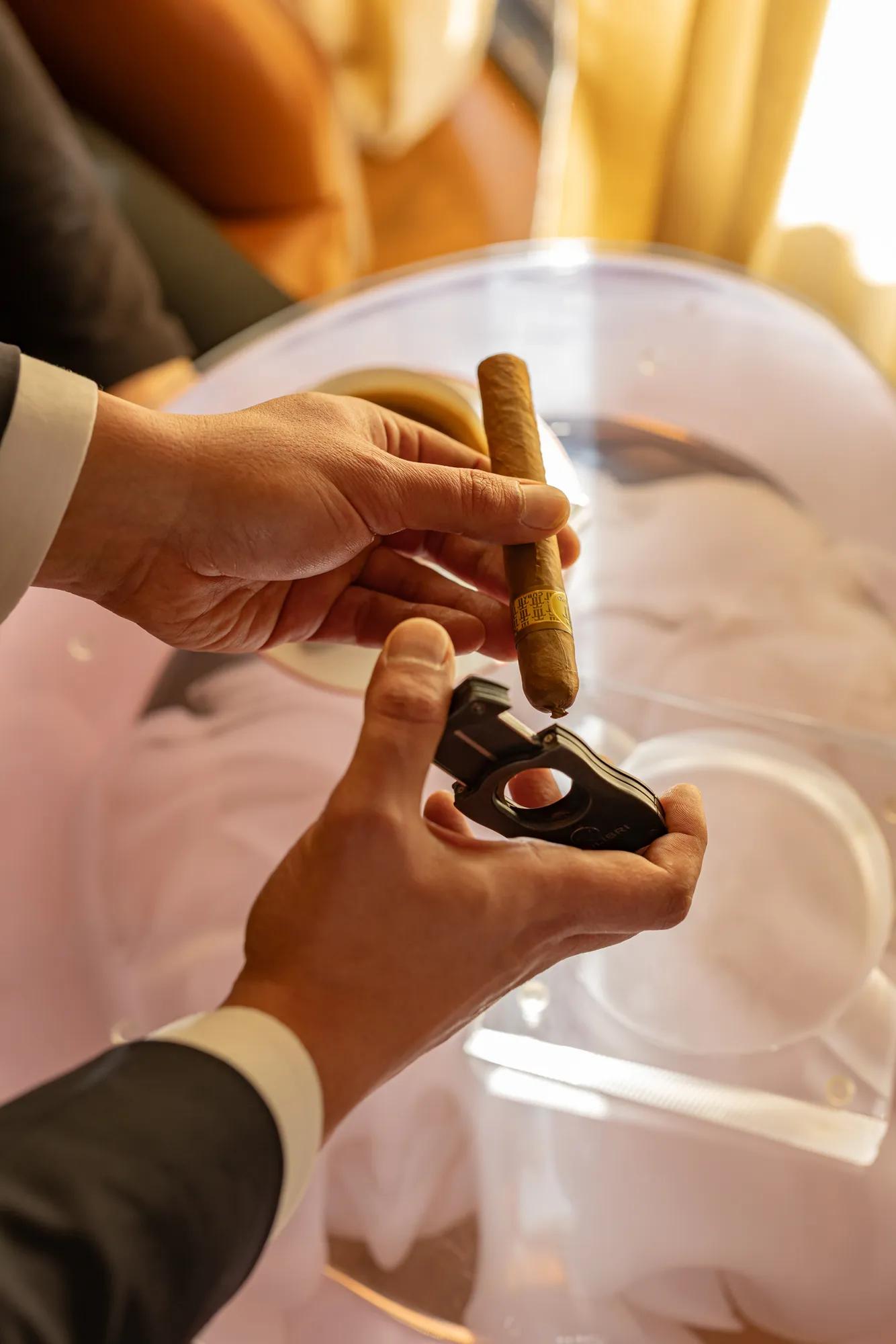 Hands holding a cigar and cutter over a glass table, preparing to cut the tip in warm light