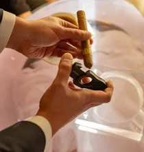 Hands holding a cigar and cutter over a glass table, preparing to cut the tip in warm light