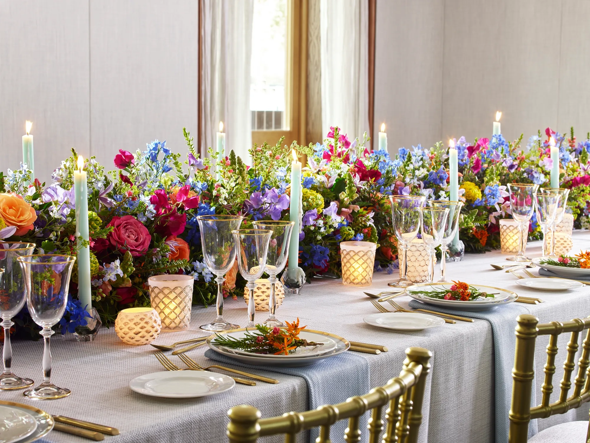 Elegant banquet table set with gold chairs, glassware and candles, decorated with vibrant floral arrangements and pastel taper candles.