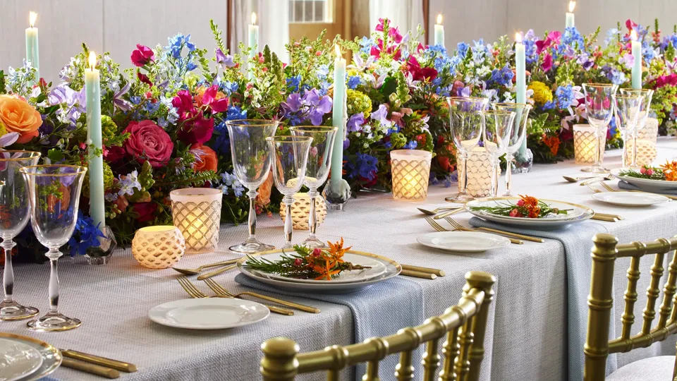 Elegant banquet table set with gold chairs, glassware and candles, decorated with vibrant floral arrangements and pastel taper candles.
