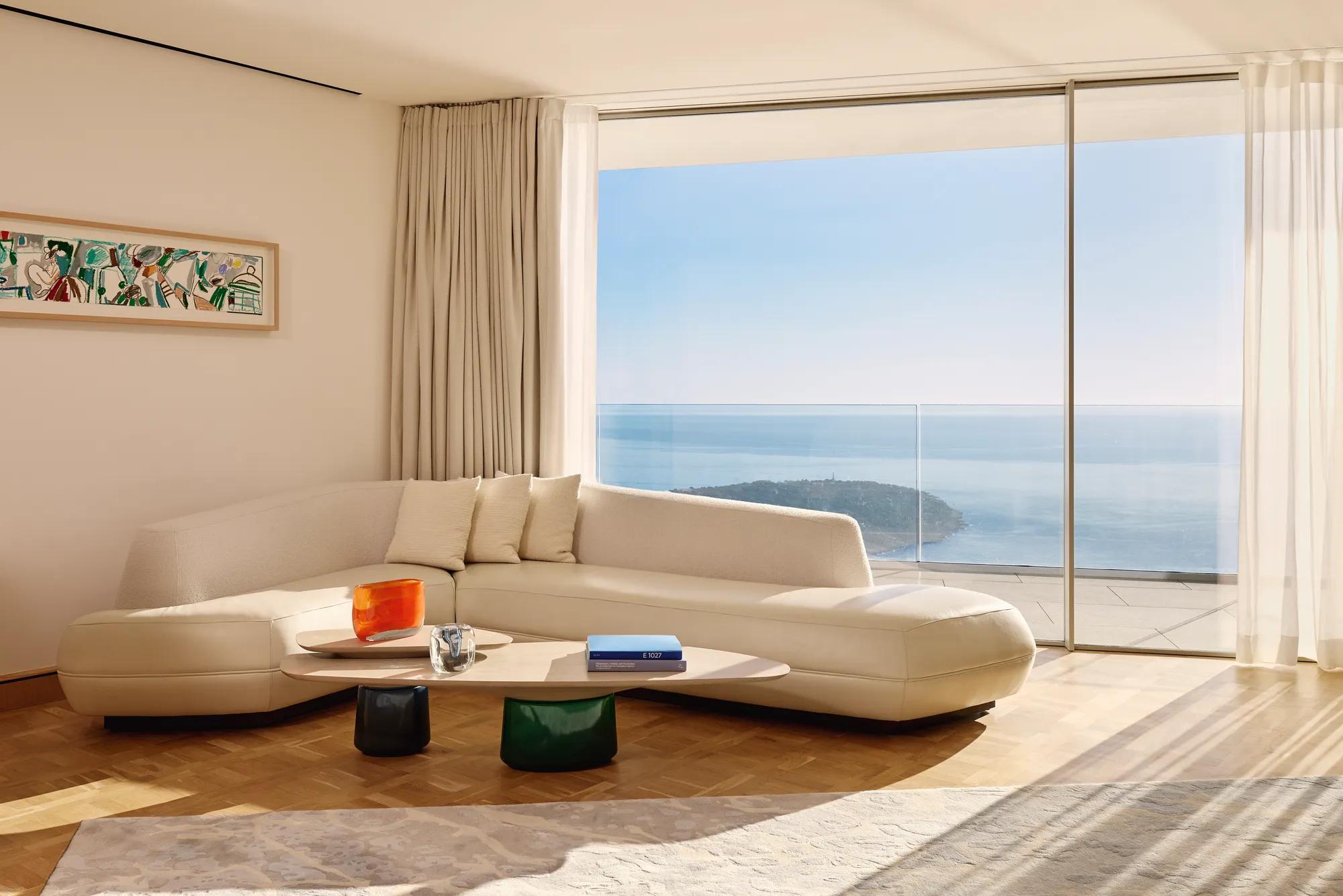 Salon moderne avec un canapé incurvé de couleur crème et une table basse en bois, placé devant de grandes baies vitrées offrant une vue panoramique sur l’océan et le littoral. La pièce est baignée de lumière naturelle, décorée avec un mobilier minimaliste, des rideaux légers et une œuvre d’art abstraite encadrée au mur.