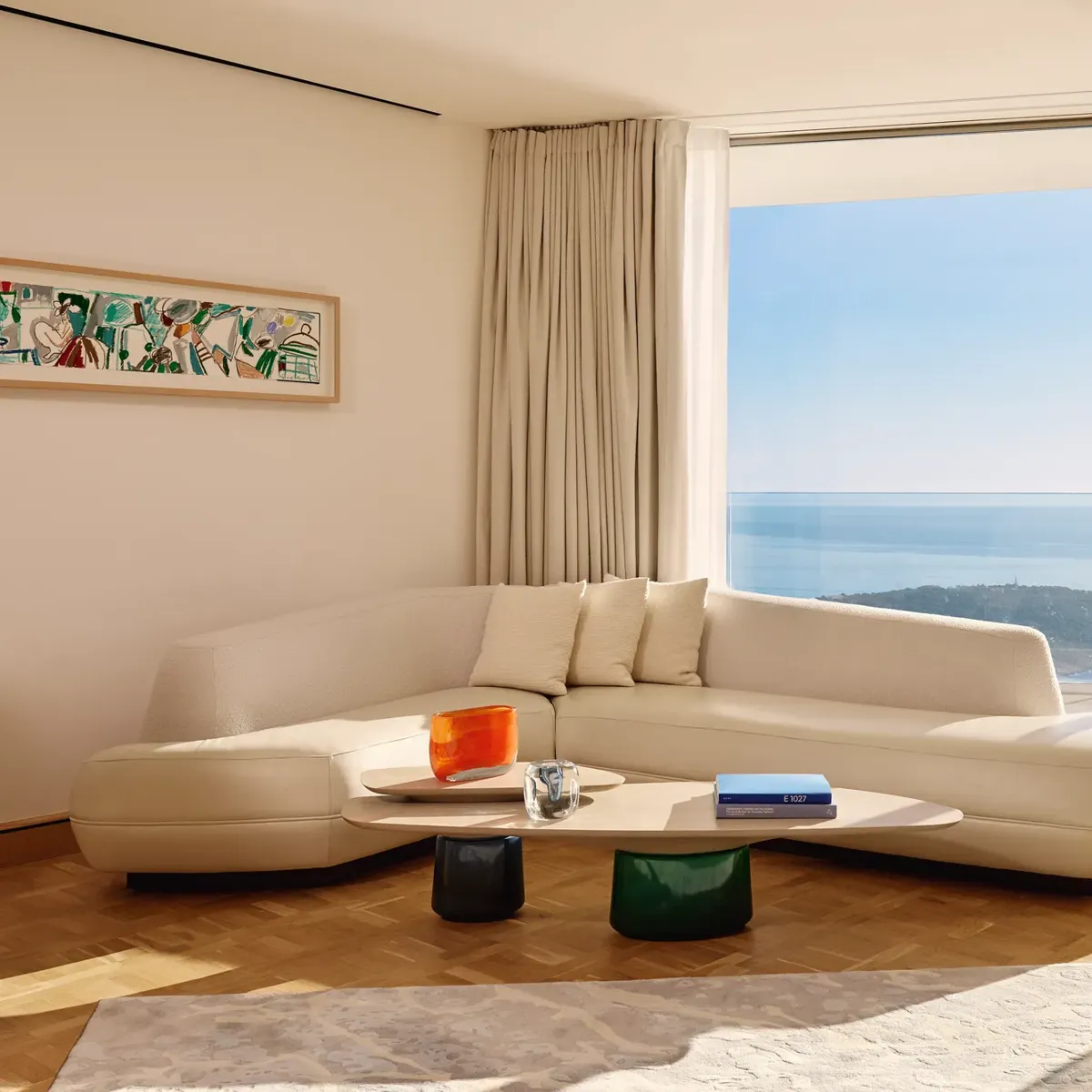Salon moderne avec un canapé incurvé de couleur crème et une table basse en bois, placé devant de grandes baies vitrées offrant une vue panoramique sur l’océan et le littoral. La pièce est baignée de lumière naturelle, décorée avec un mobilier minimaliste, des rideaux légers et une œuvre d’art abstraite encadrée au mur.