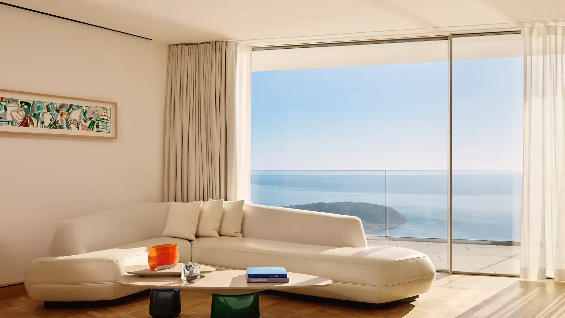 Modern living room with a curved cream-colored sofa and low wooden coffee table, set against floor-to-ceiling windows offering a panoramic view of the ocean and coastline. Soft natural light fills the room, which is decorated with minimal furniture, sheer curtains, and a framed abstract artwork on the wall.