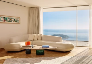 Salon moderne avec un canapé incurvé de couleur crème et une table basse en bois, placé devant de grandes baies vitrées offrant une vue panoramique sur l’océan et le littoral. La pièce est baignée de lumière naturelle, décorée avec un mobilier minimaliste, des rideaux légers et une œuvre d’art abstraite encadrée au mur.