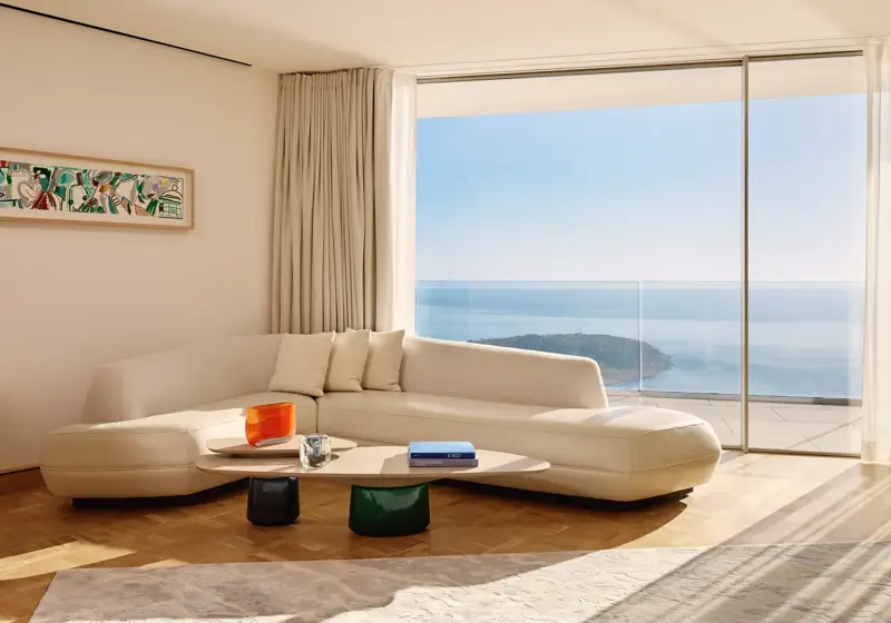 Salon moderne avec un canapé incurvé de couleur crème et une table basse en bois, placé devant de grandes baies vitrées offrant une vue panoramique sur l’océan et le littoral. La pièce est baignée de lumière naturelle, décorée avec un mobilier minimaliste, des rideaux légers et une œuvre d’art abstraite encadrée au mur.