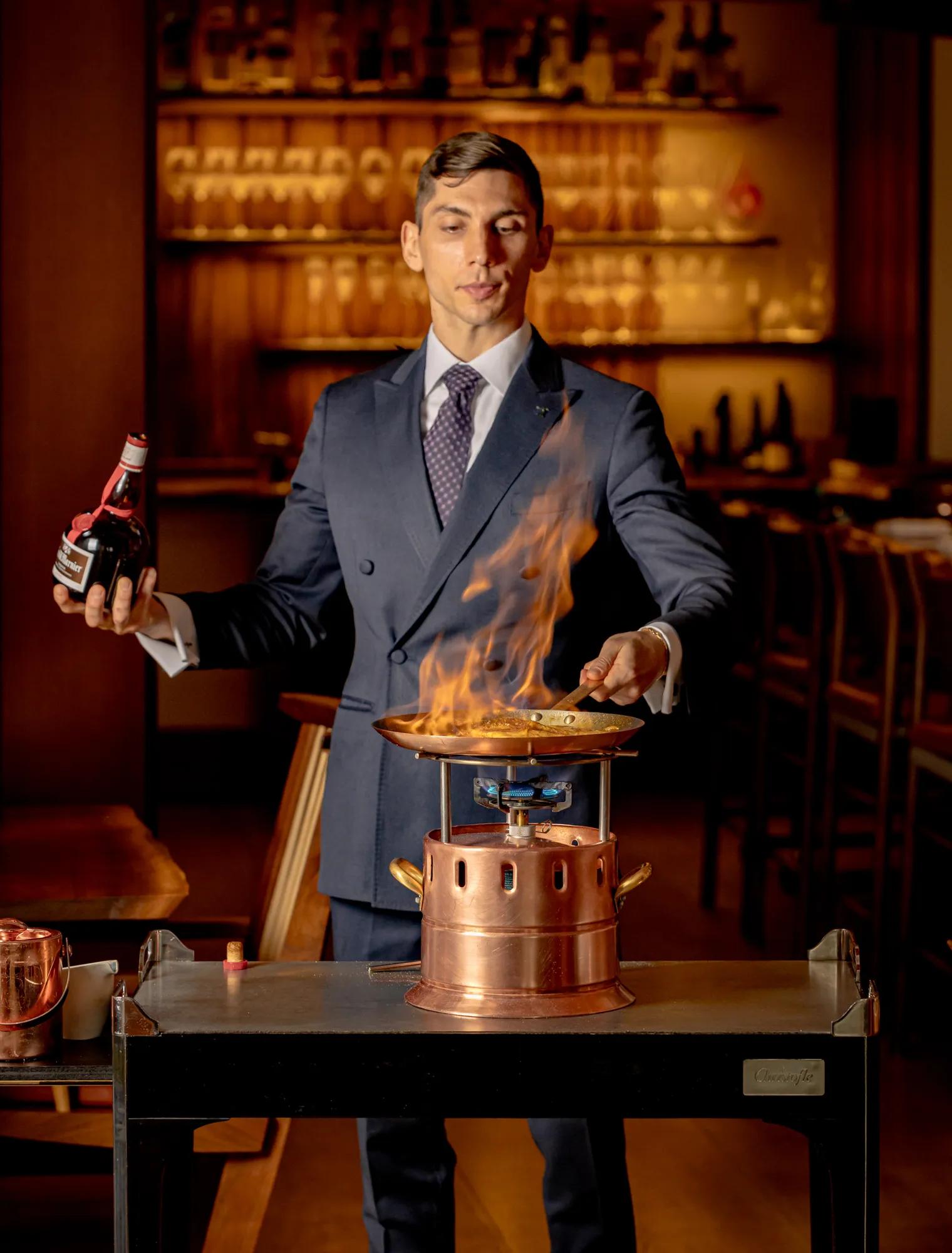 Restaurant staff member preparing a flaming dish at a tableside serving trolley.