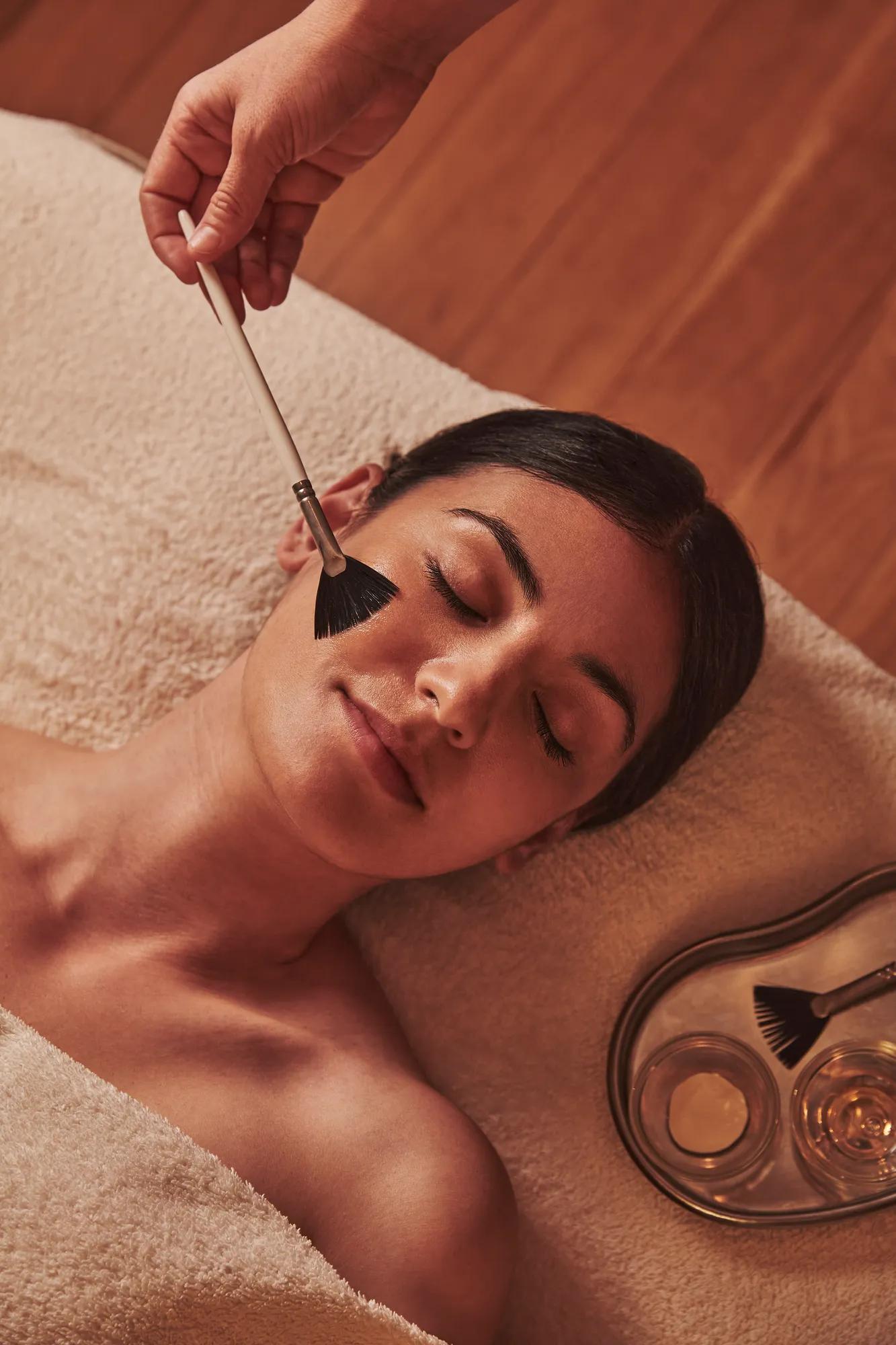 Spa therapist applying a facial treatment with a soft brush to a woman lying relaxed on a towel.