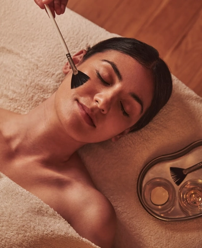Spa therapist applying a facial treatment with a soft brush to a woman lying relaxed on a towel.