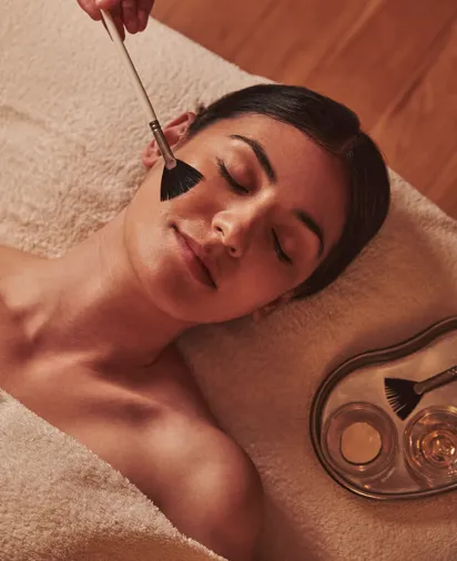 Spa therapist applying a facial treatment with a soft brush to a woman lying relaxed on a towel.